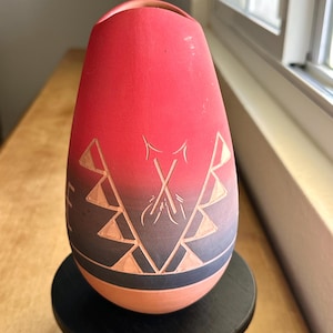May include: A red and gray ceramic vase with etched geometric designs and crossed arrows. The vase has a rounded shape and sits on a small black stand. The design includes triangles and a gradient effect.