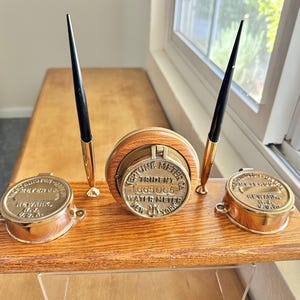 Puede incluir: Un juego de escritorio de madera con dos tinteros de latón y un contador de agua de latón. El contador de agua indica "Neptune Meter Co. Trident 665065 Water Meter New York".