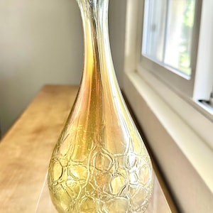 Puede incluir: Un jarrón de vidrio transparente con un patrón texturizado similar a burbujas en la mitad inferior. El jarrón tiene un cuello delgado y una parte superior acampanada en forma de pétalo. El vidrio tiene un brillo dorado e iridiscente.