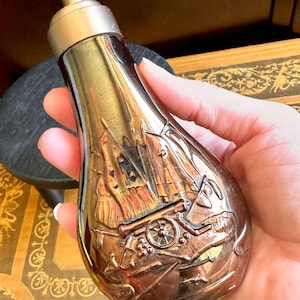 May include: A metallic brown glass powder flask with a teardrop shape and a silver-toned metal spout. The surface features a raised relief design depicting a sailing ship, a cannon, and various nautical elements, finished with a reflective sheen.