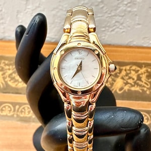 May include: A gold-toned Seiko wristwatch with a white face and a small diamond accent at the 12 o'clock position. The watch has a gold band and a round face. The watch is displayed on a black hand-shaped stand.