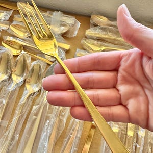 May include: A gold-colored fork with a long, slender handle. The fork is held in a hand, and there are many other gold-colored forks and spoons in the background.