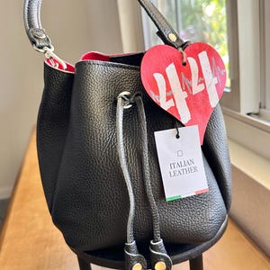 May include: Black leather bucket bag with a red interior. The bag features a drawstring closure and a top handle. A heart-shaped tag with the number 4424 is attached, along with a tag that says "ITALIAN LEATHER".