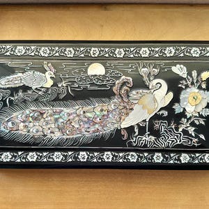 Può includere: Scatola rettangolare nera con un design intarsiato. Il design presenta un pavone con piume iridescenti, un uccello bianco, un sole ed elementi floreali. La scatola è incorniciata da un bordo floreale bianco.
