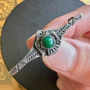 May include: A silver brooch featuring a central round green stone set within a bird-shaped frame. The elongated wings of the bird are decorated with etched linear patterns and geometric triangles, creating a detailed metallic surface texture.