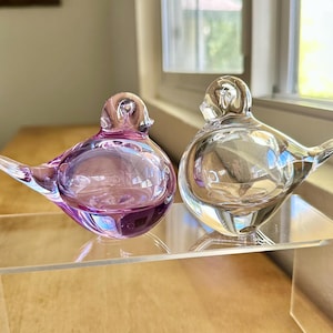 May include: Two glass bird figurines. One is clear, the other is a gradient of pink and purple. Both have rounded bodies, pointed beaks, and outstretched wings. They sit on a clear acrylic display stand.