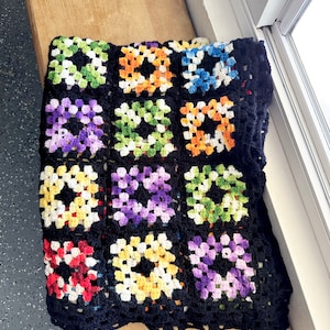 Puede incluir: Una manta de crochet negra con cuadrados de granny coloridos. Los cuadrados están hechos en una variedad de colores, incluyendo rojo, naranja, amarillo, verde, azul y morado.