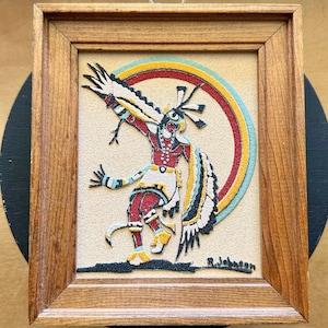 Puede incluir: Una obra de arte en arena enmarcada que presenta una figura estilizada en una pose dinámica sobre un fondo circular multicolor de rojo, amarillo y azul claro. La figura lleva adornos detallados en blanco, rojo y negro. Firmado R. Johnson.