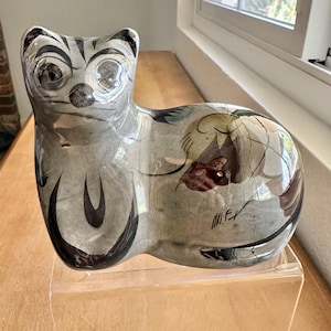 Puede incluir: Figura de gato de cerámica en tonos grises, negros y marrones. El gato tiene ojos grandes y expresivos y un diseño estilizado. La figura descansa sobre un soporte de acrílico transparente, con una firma en el lateral.