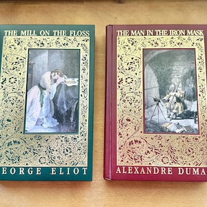 May include: Two hardcover books: "The Mill on the Floss" by George Eliot and "The Man in the Iron Mask" by Alexandre Dumas. The books have decorative gold and green or red covers with illustrations. The titles and authors are printed on the covers.