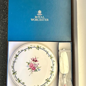 May include: A Royal Worcester porcelain cake plate and matching server set in a blue presentation box. The white plate features a central floral bouquet, a gold rim, and a blue border with green vines. The server has a floral handle.