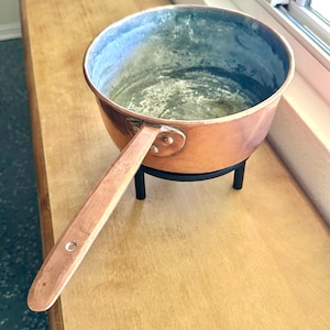 May include: A copper saucepan with a long handle, resting on a black metal stand. The interior of the pan shows signs of use. The pan is placed on a wooden surface near a window.