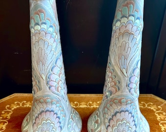 May include: A pair of ceramic candle holders featuring a tapered cylindrical shape with a flared base and top. The surface is decorated with a repeating fan-like pattern in muted shades of blue, pink, and grey with gold-toned rim accents.