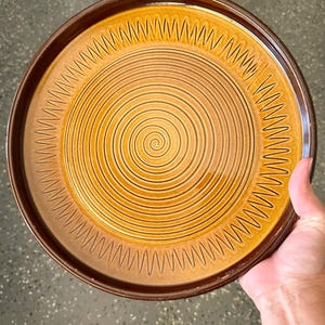 Puede incluir: Un plato redondo de cerámica con un diseño en espiral en tonos marrones y amarillos. El plato tiene un borde marrón oscuro y una serie de patrones en zigzag alrededor del borde. El plato es sostenido por una mano.