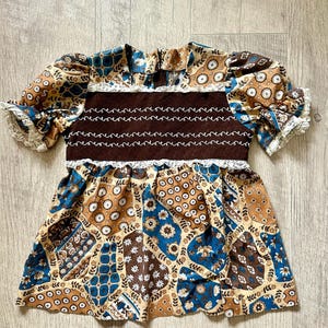 Puede incluir: Vestido de estilo vintage con diseño de patchwork en tonos marrón, azul y beige. Tiene mangas cortas abullonadas con ribete de encaje y un corpiño marrón oscuro con detalles bordados en blanco. La falda acampanada evoca los años 70.