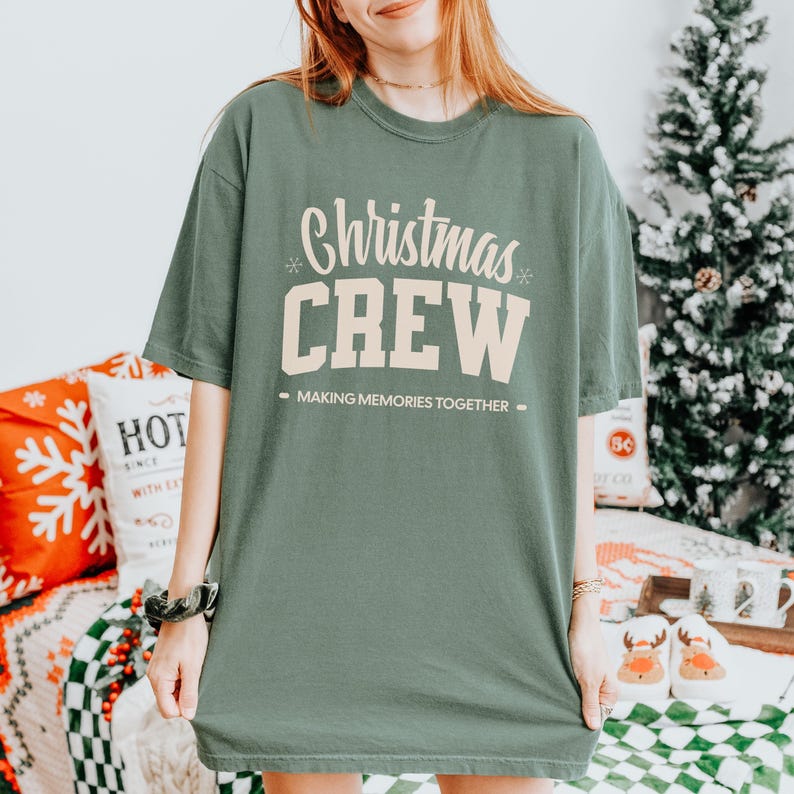 May include: A sage green t-shirt with the words "Christmas CREW" in a cream-coloured, stylised font. The text "MAKING MEMORIES TOGETHER" is below the word "CREW". The shirt is a relaxed fit and has short sleeves.