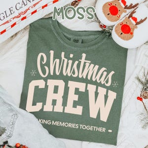 May include: A moss green t-shirt with the words "Christmas CREW" in cream. The text "MAKING MEMORIES TOGETHER" is below. The image also shows reindeer slippers and a decorative pillow.