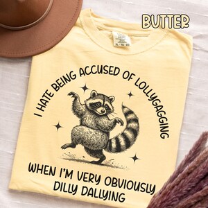 May include: Butter-colored t-shirt featuring a dancing raccoon illustration. The text reads "I HATE BEING ACCUSED OF LOLLYGAGGING WHEN I'M VERY OBVIOUSLY DILLY DALLYING." The shirt is a comfortable XL size.