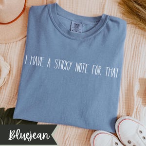 May include: A bluejean-colored t-shirt with the text "I HAVE A STICKY NOTE FOR THAT" in white. The shirt has a crew neck and short sleeves. The Comfort Colors brand tag is visible. White sneakers are in the bottom right corner.