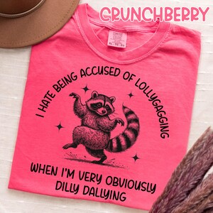 May include: A bright pink t-shirt with the word "CRUNCHBERRY" in pink script. The shirt features a raccoon graphic with the text "I HATE BEING ACCUSED OF LOLLYGAGGING WHEN I'M VERY OBVIOUSLY DILLY DALLYING."