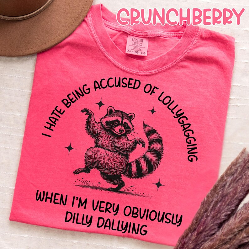 May include: A bright pink t-shirt with the word "CRUNCHBERRY" in pink script. The shirt features a raccoon graphic with the text "I HATE BEING ACCUSED OF LOLLYGAGGING WHEN I'M VERY OBVIOUSLY DILLY DALLYING."