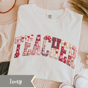 May include: Ivory t-shirt featuring the word "TEACHER" appliqu&eacute;d across the front. The letters are crafted from diverse red and pink patterned fabrics, including hearts and plaid. The shirt is a Comfort Colors brand.