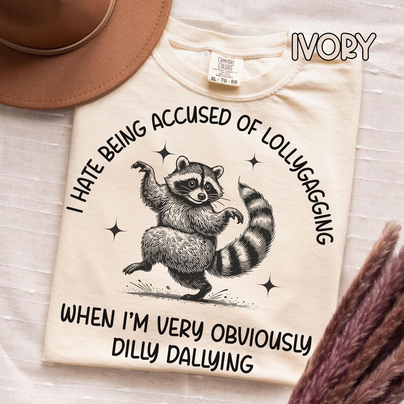 May include: Ivory-colored t-shirt featuring a raccoon illustration. The raccoon is depicted in a playful pose with a striped tail. Text on the shirt reads, "I HATE BEING ACCUSED OF LOLLYGAGGING WHEN I'M VERY OBVIOUSLY DILLY DALLYING."