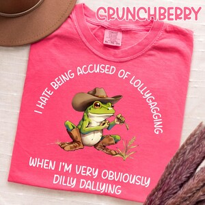 May include: A pink t-shirt with the word "CRUNCHBERRY" at the top. The shirt features a cartoon frog wearing a cowboy hat and boots, holding a piece of grass. Text around the frog reads "I HATE BEING ACCUSED OF LOLLYGAGGING WHEN I'M VERY OBVIOUSLY DILLY DALLYING."