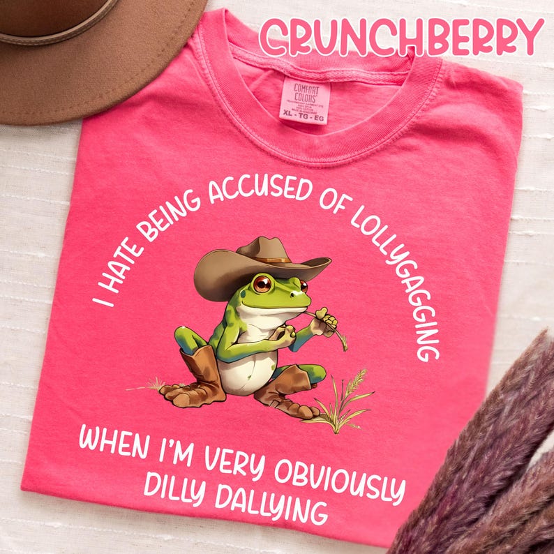 May include: A pink t-shirt with the word "CRUNCHBERRY" at the top. The shirt features a cartoon frog wearing a cowboy hat and boots, holding a piece of grass. Text around the frog reads "I HATE BEING ACCUSED OF LOLLYGAGGING WHEN I'M VERY OBVIOUSLY DILLY DALLYING."