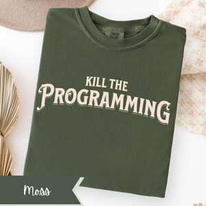May include: A moss green t-shirt with the text "KILL THE PROGRAMMING" in a vintage-style font. The shirt is folded and displayed on a white surface, with a neutral-colored hat and decorative fabric in the background. The word "Moss" is displayed on a banner.