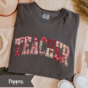 May include: A dark grey t-shirt with the word "TEACHER" appliqu&eacute;d across the front in a patchwork design. The patchwork includes red hearts and plaid patterns. The shirt is a Comfort Colors brand.