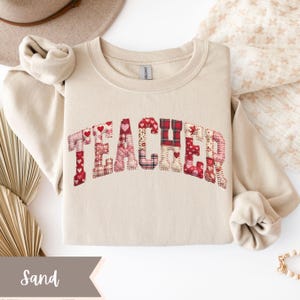 May include: A sand-colored sweatshirt with the word "TEACHER" appliquéd across the chest. The letters are made of patchwork fabric with red hearts and plaid patterns. The sweatshirt is folded neatly.