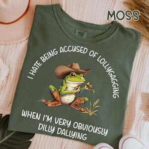 May include: Moss green t-shirt with a cartoon frog wearing a cowboy hat and boots. The frog is holding a piece of straw. Text around the frog says, "I hate being accused of lollygagging when I'm very obviously dilly dallying."