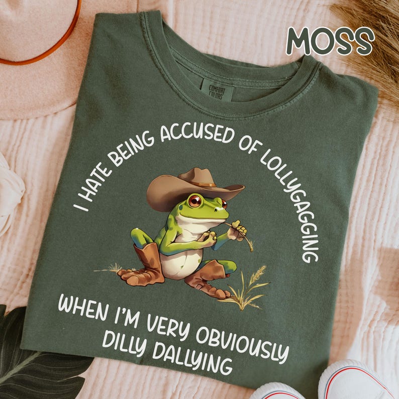 May include: Moss green t-shirt with a cartoon frog wearing a cowboy hat and boots. The frog is holding a piece of straw. Text around the frog says, "I hate being accused of lollygagging when I'm very obviously dilly dallying."