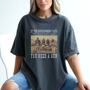 May include: Dark gray t-shirt with the text "IF THE GOVERNMENT SAYS 'You don't need a gun' YOU NEED A GUN" above a sepia-toned image of people holding rifles. The shirt is a relaxed fit.
