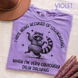 May include: A violet t-shirt with a graphic of a raccoon dancing. The text on the shirt reads "I HATE BEING ACCUSED OF LOLLYGAGGING WHEN I'M VERY OBVIOUSLY DILLY DALLYING". The shirt is made of a soft material.