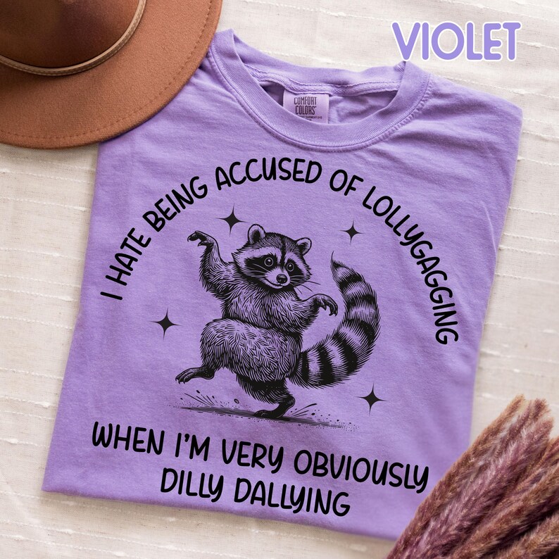 May include: A violet t-shirt with a graphic of a raccoon dancing. The text on the shirt reads "I HATE BEING ACCUSED OF LOLLYGAGGING WHEN I'M VERY OBVIOUSLY DILLY DALLYING". The shirt is made of a soft material.