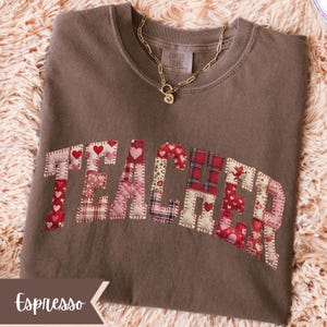 May include: Espresso-coloured sweatshirt featuring the word "TEACHER" appliqu&eacute;d across the front in a patchwork design of red and pink hearts and plaid patterns. A gold chain necklace with a pendant rests on the shirt.