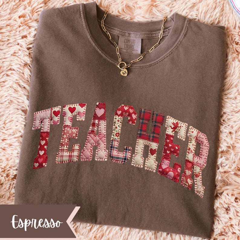 May include: Espresso-coloured sweatshirt featuring the word "TEACHER" appliqu&eacute;d across the front in a patchwork design of red and pink hearts and plaid patterns. A gold chain necklace with a pendant rests on the shirt.