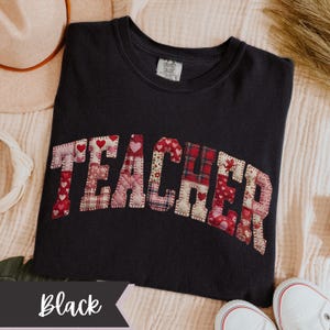May include: Black t-shirt featuring the word "TEACHER" in patchwork lettering. The letters are made from fabric with heart and floral designs. The shirt is a Comfort Colors brand.