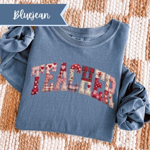 May include: A bluejean-coloured long-sleeved shirt with the word "TEACHER" appliquéd across the front. The letters are made of patchwork fabric with heart and plaid patterns. The shirt is folded on a woven surface.