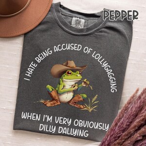 May include: Dark gray t-shirt with a graphic of a frog wearing a cowboy hat and boots, holding a piece of grass. The text on the shirt reads "I HATE BEING ACCUSED OF LOLLYGAGGING WHEN I'M VERY OBVIOUSLY DILLY DALLYING."