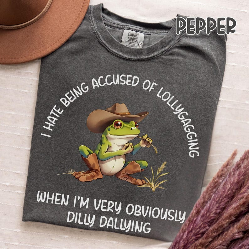 May include: Dark gray t-shirt with a graphic of a frog wearing a cowboy hat and boots, holding a piece of grass. The text on the shirt reads "I HATE BEING ACCUSED OF LOLLYGAGGING WHEN I'M VERY OBVIOUSLY DILLY DALLYING."