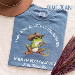May include: Blue jean t-shirt with a cartoon frog wearing a cowboy hat and boots. The frog is holding a piece of grass. Text on the shirt reads "I HATE BEING ACCUSED OF LOLLYGAGGING WHEN I'M VERY OBVIOUSLY DILLY DALLYING."