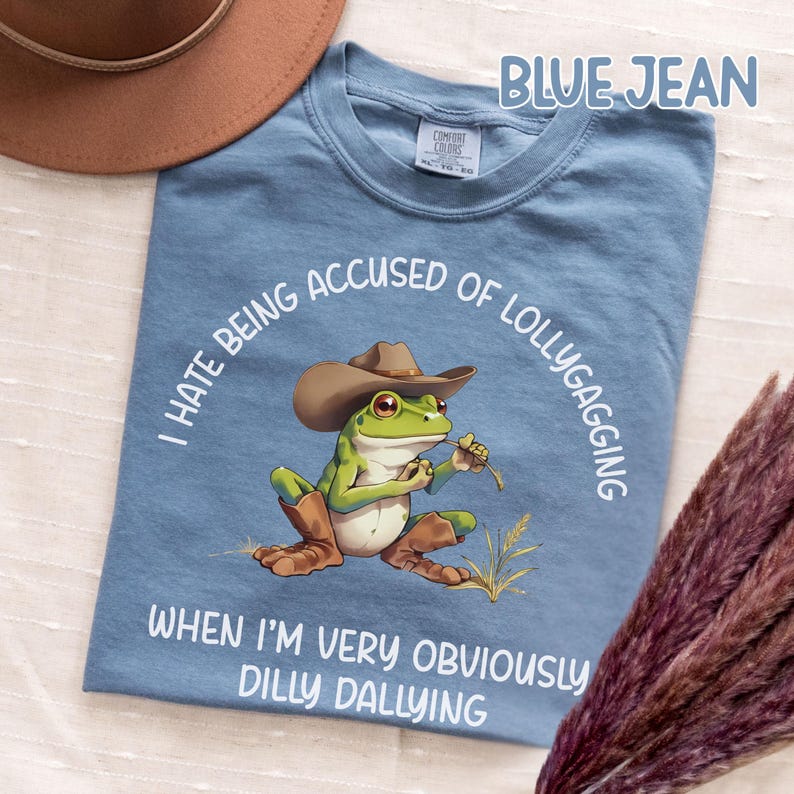 May include: Blue jean t-shirt with a cartoon frog wearing a cowboy hat and boots. The frog is holding a piece of grass. Text on the shirt reads "I HATE BEING ACCUSED OF LOLLYGAGGING WHEN I'M VERY OBVIOUSLY DILLY DALLYING."