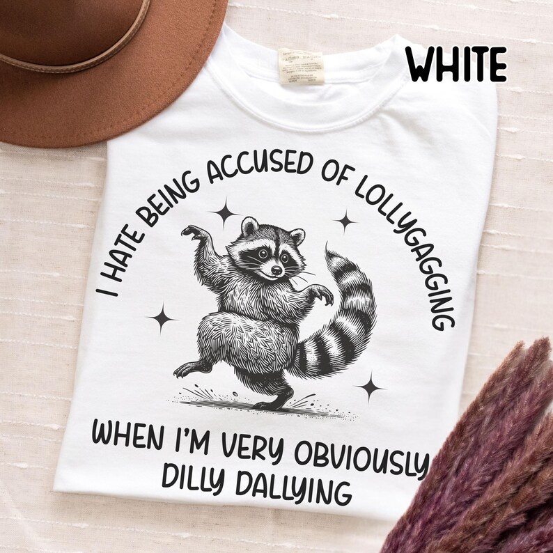 May include: White t-shirt with a raccoon graphic and the text "I hate being accused of lollygagging when I'm very obviously dilly dallying." The raccoon is black and white with a striped tail. The word "WHITE" is printed at the top.