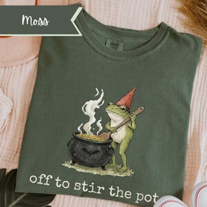 May include: A moss green t-shirt featuring a graphic of a green frog wearing a red polka-dot party hat and stirring a black cauldron with a wooden spoon. White steam rises from the pot, which is decorated with a bat and spiderweb. Text reads: off to stir the pot.