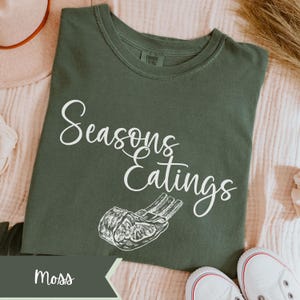 May include: A moss green t-shirt with the words "Seasons Eatings" in white script, along with a graphic of a rack of ribs. The shirt is made of a soft material and has a crew neck. The word "Moss" is in a banner at the bottom.