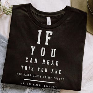 May include: Black t-shirt with white text that reads "IF YOU CAN READ THIS YOU ARE TOO DAMN CLOSE TO MY COFFEE ARE YOU BLIND? BACK OFF!". The shirt is folded and resting on a white surface.