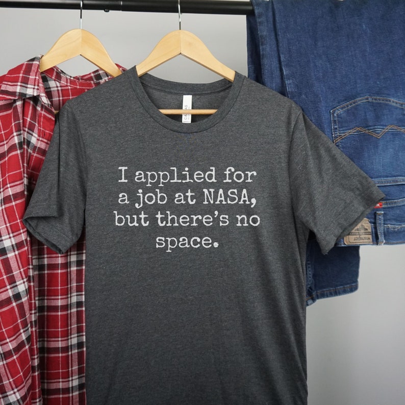 May include: A dark gray t-shirt with the text "I applied for a job at NASA, but there's no space." printed in white.